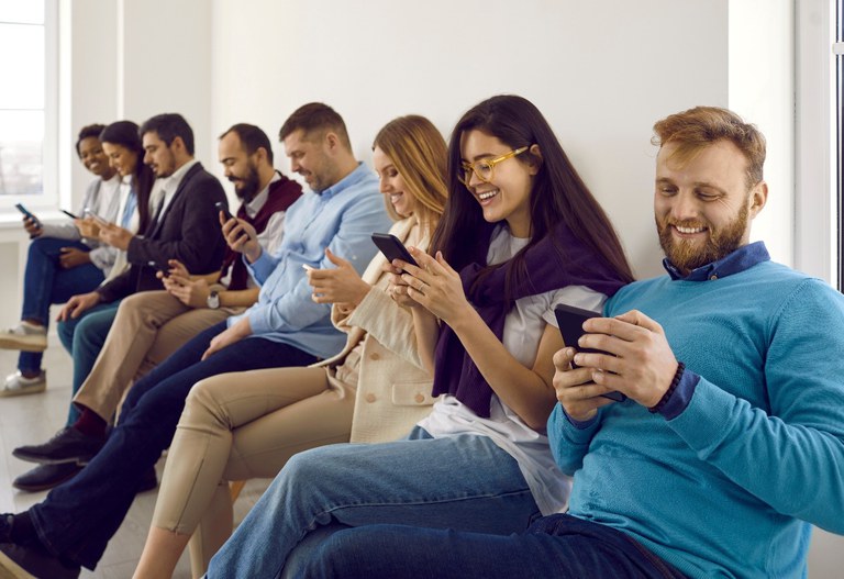 Glückliche moderne Menschen sitzen in einer Reihe zusammen und schauen auf die Bildschirme ihrer Mobiltelefone.